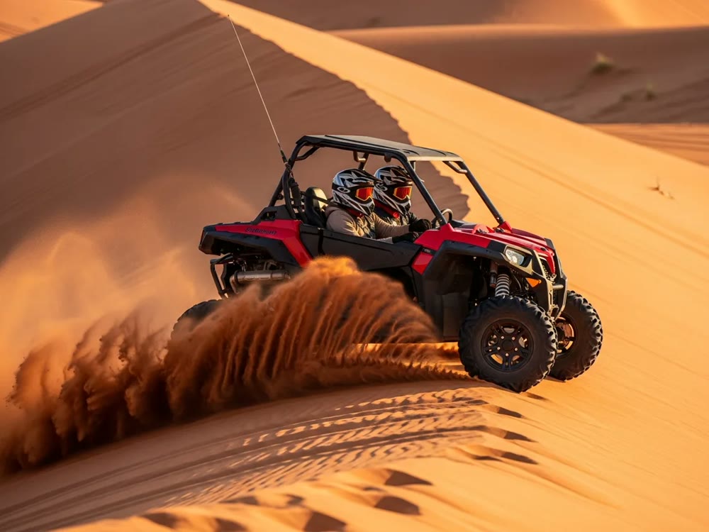 Two helmeted riders driving a red 2 seater dune buggy across a Dubai dune with sand spraying behind